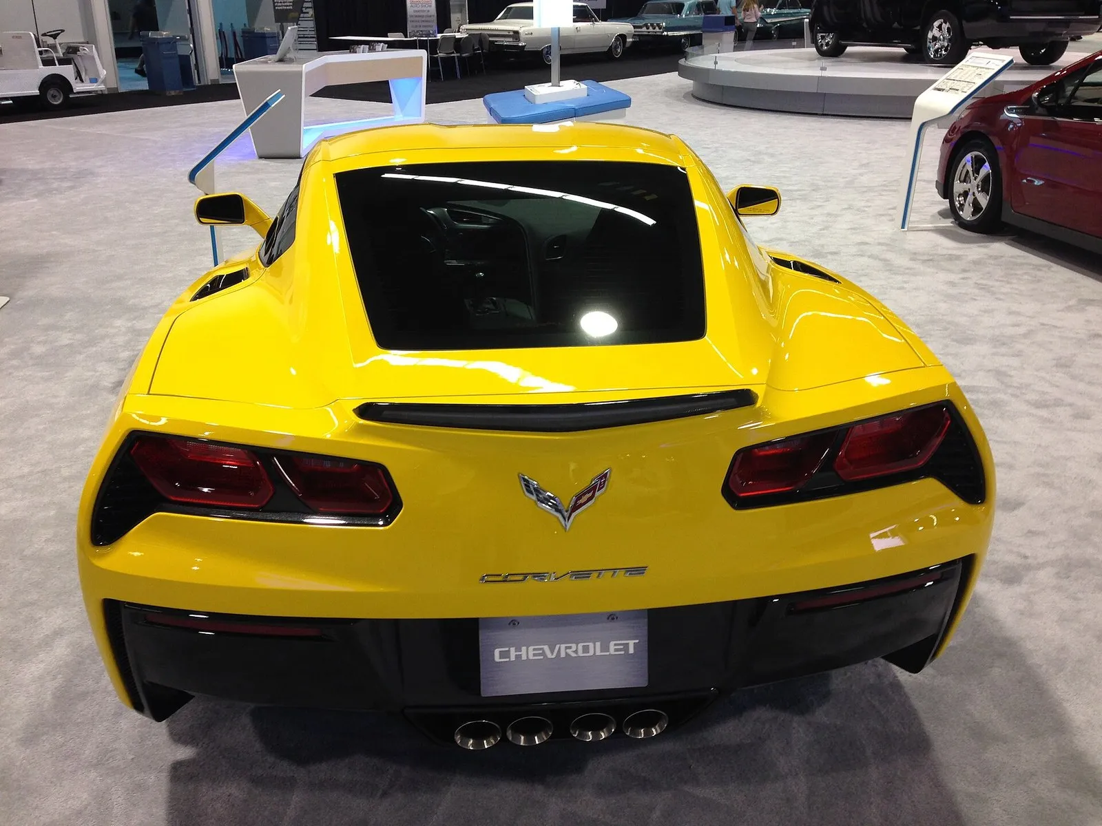 Yellow C7 Corvette Stingray coupe seen from above and behind at an auto show, showing the glass rear hatch over the cargo area.