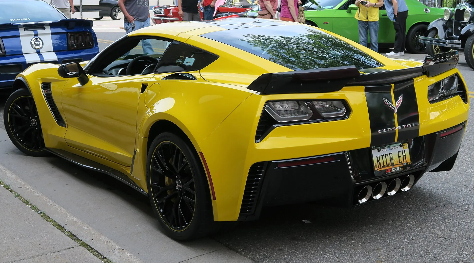 Yellow 2015 C7 Corvette Z06 coupe seen from the rear left corner at a car show, showing its wide rear fenders, a raised rear spoiler and four center exhaust tips.