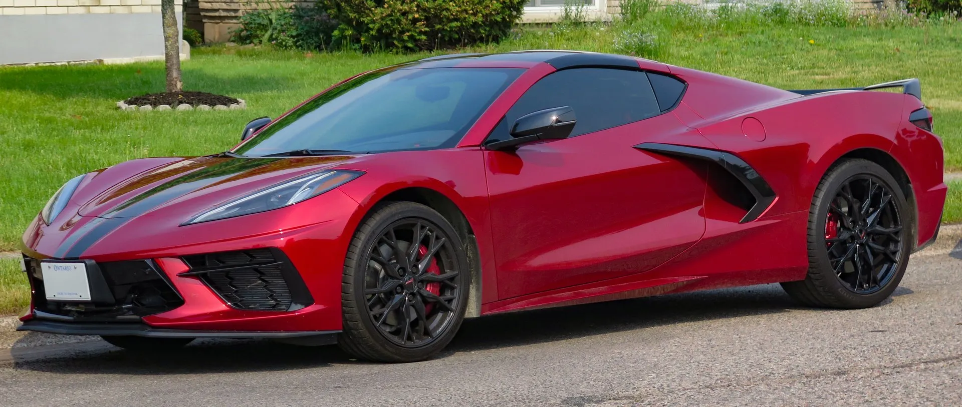 A red C8 Corvette Stingray coupe with a dark stripe over its nose, black wheels and red brake calipers, parked at a curb in front of a lawn, seen from the front left.