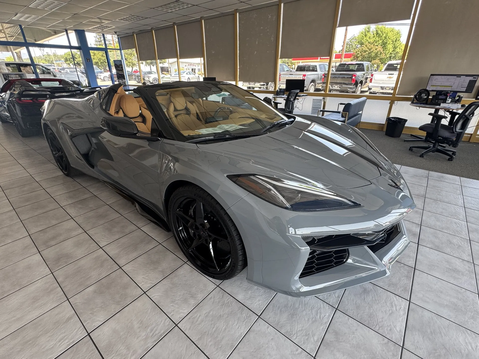 A gray C8 Corvette E-Ray convertible with its top stowed, a tan interior and gloss black wheels, on the tiled floor of a dealer's showroom, with desks inside and pickup trucks on the lot behind.