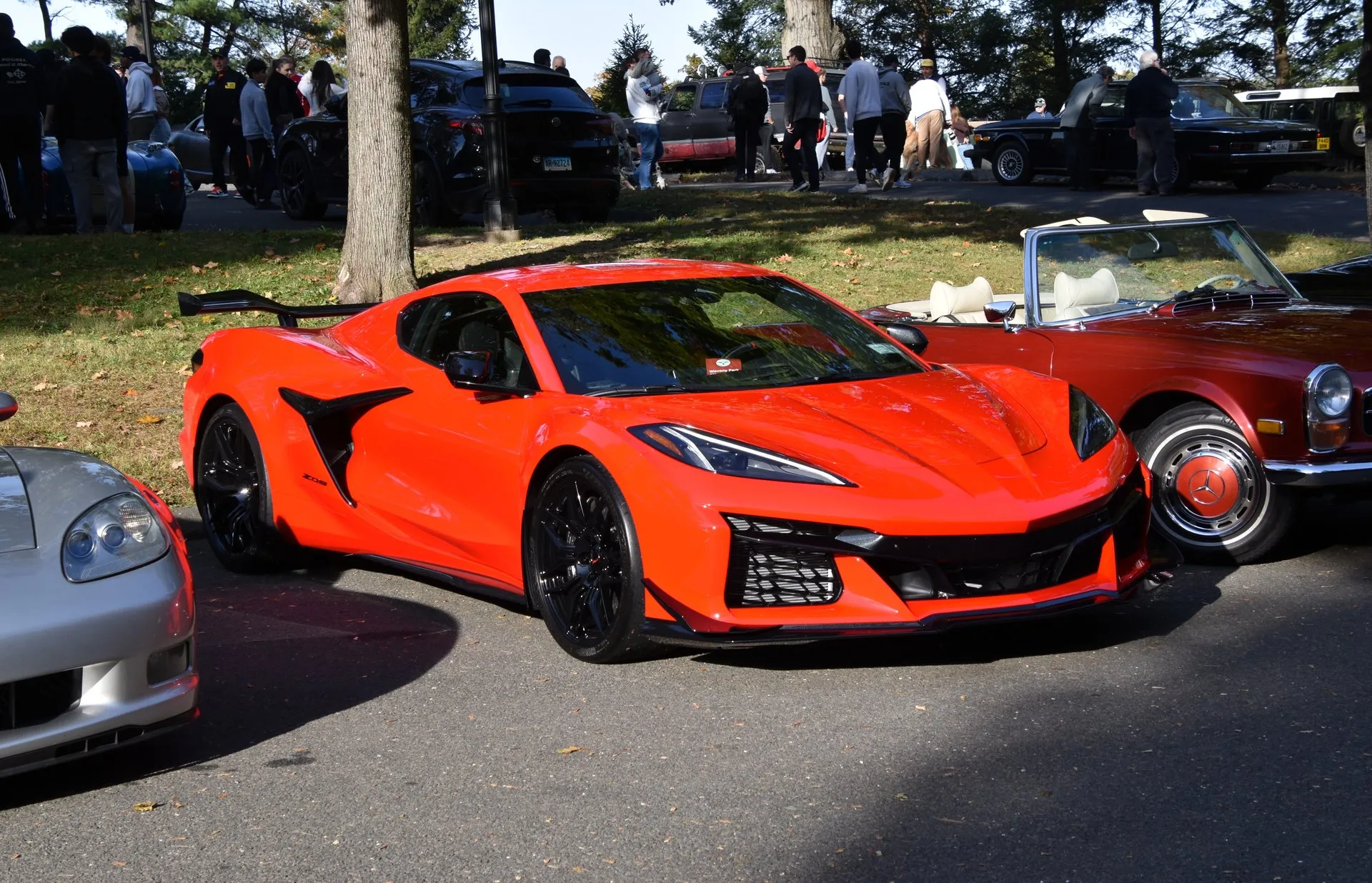 An orange 2025 C8 Corvette Z06 coupe with black wheels, black side air intakes and a tall rear wing, parked at a car show between a silver C6 Corvette and a red classic Mercedes-Benz convertible, seen from the front right.