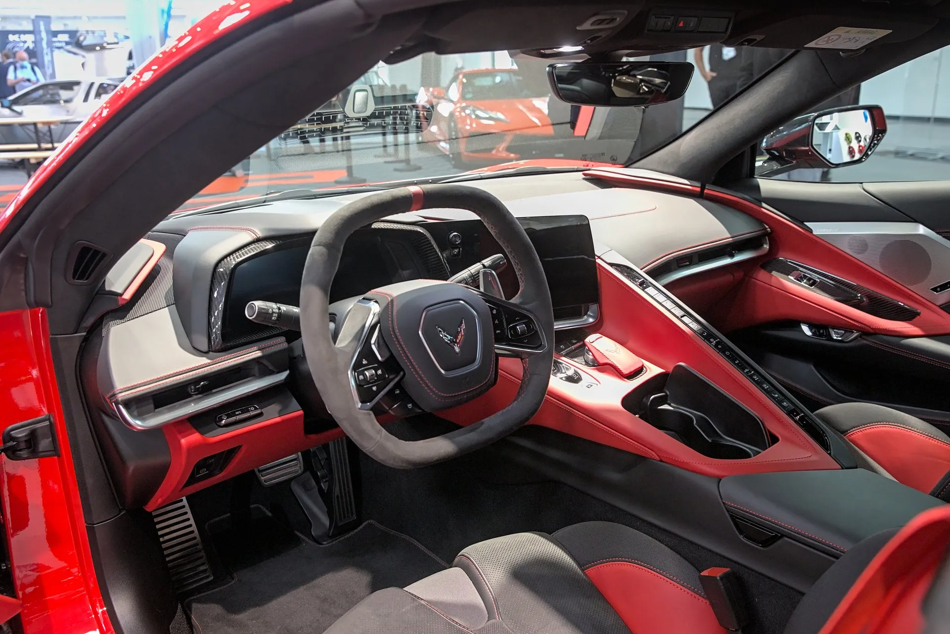 The red and black cockpit of a C8 Corvette seen through the open driver's door: a squared-off steering wheel, a digital instrument display, a center touch screen, a long row of buttons along the console and two pedals in the footwell.