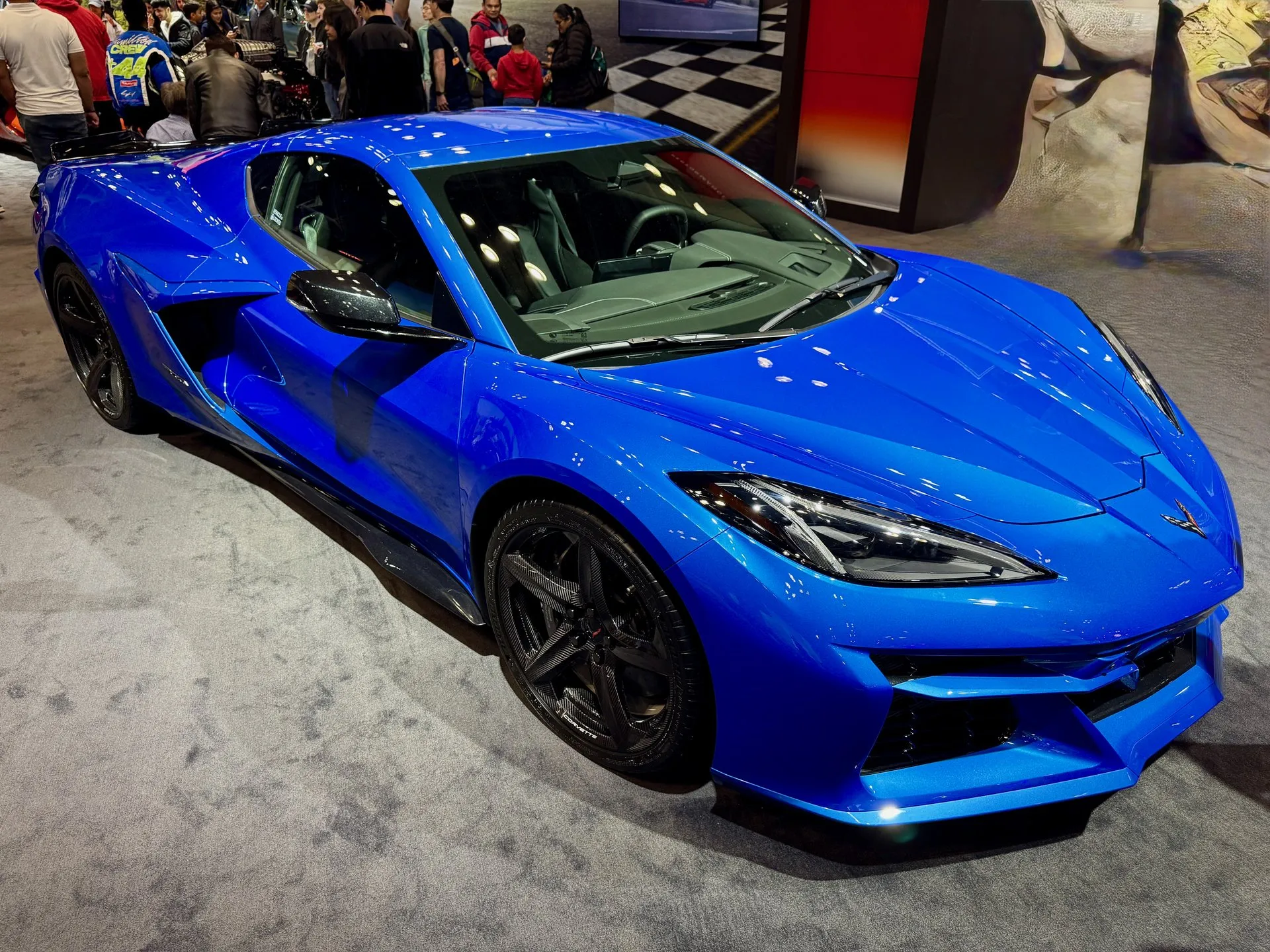 A bright blue C8 Corvette E-Ray coupe on auto show carpet, seen from above its front corner, with black carbon-weave five-spoke wheels, dark side skirts and wide rear fenders, and show visitors behind it.