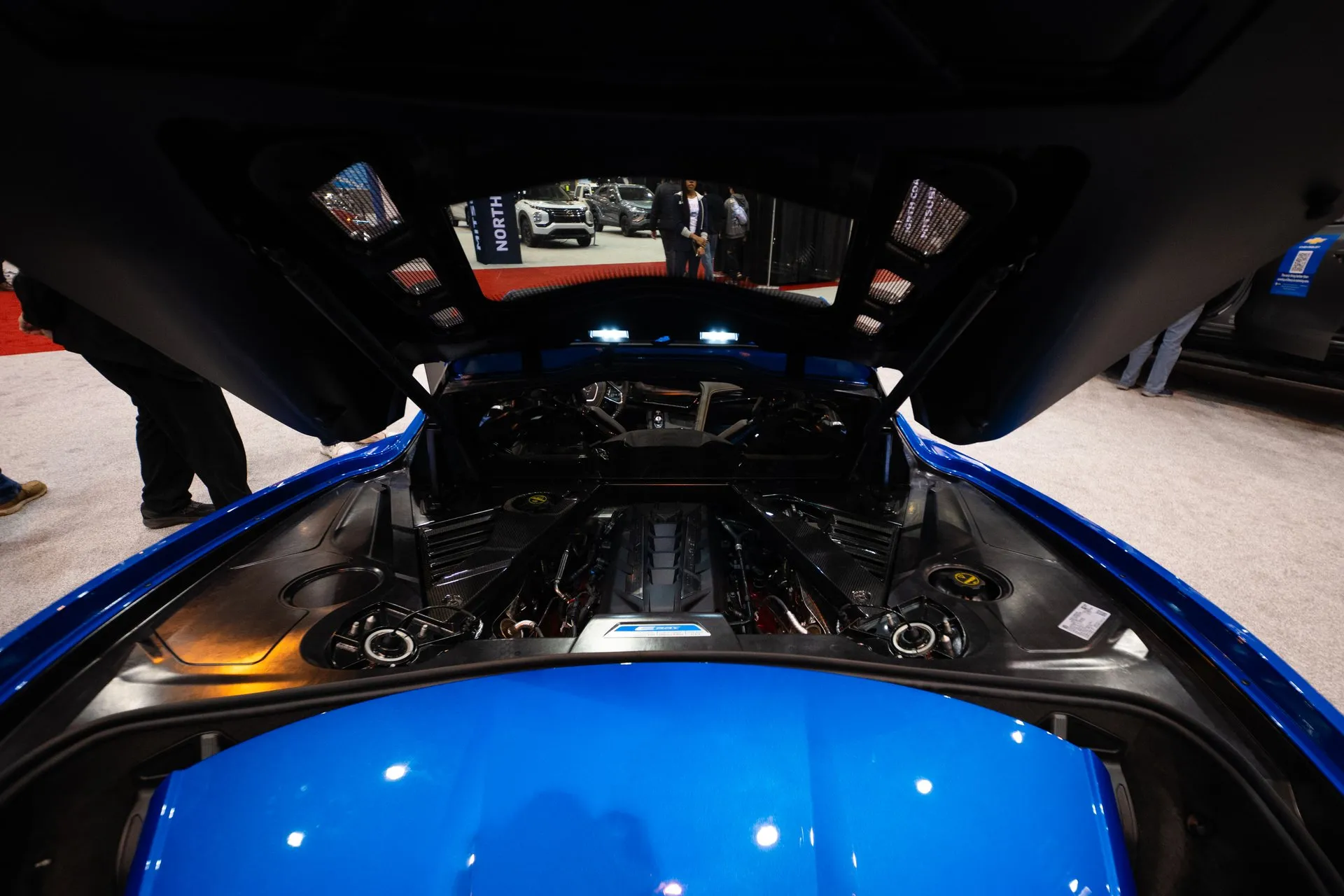The engine bay of a blue C8 Corvette E-Ray at an auto show, seen over the rear deck with the glass hatch raised: the V8 under black covers with a blue and white plaque at the near end, and the cabin visible through the rear window beyond.