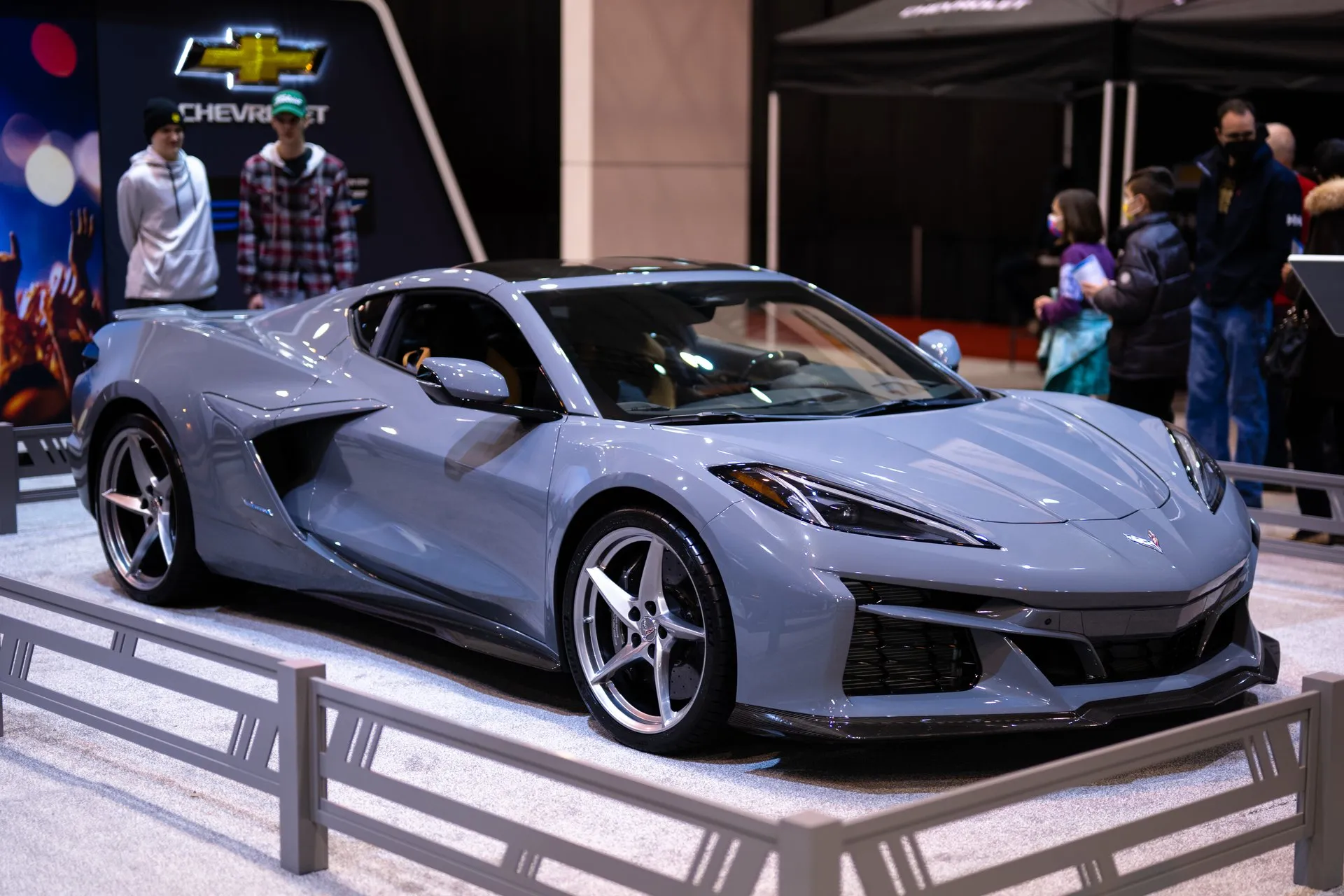 A gray C8 Corvette E-Ray coupe with silver five-spoke wheels on a fenced auto show stand, seen from the front right, with visitors and a Chevrolet sign behind it.