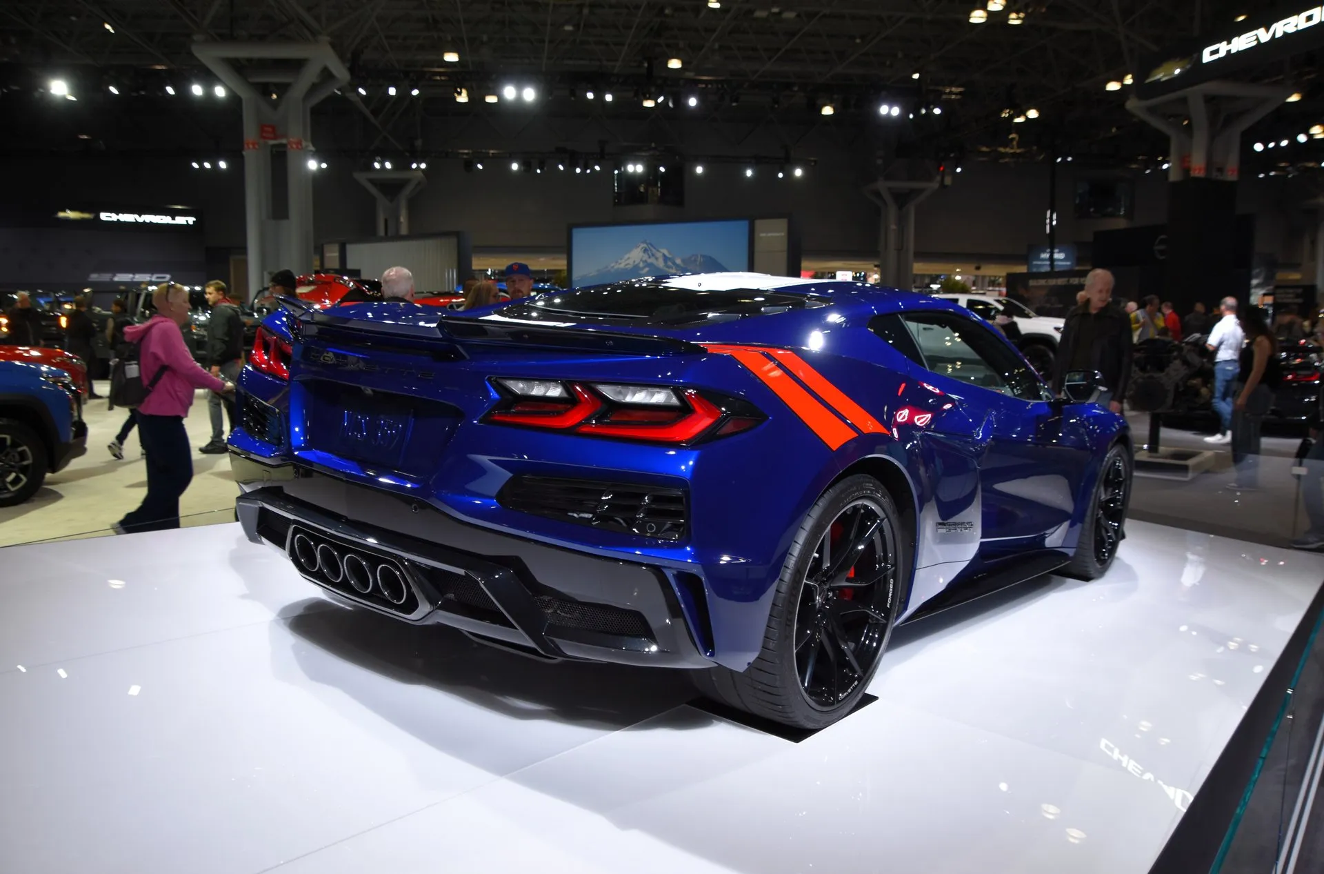 The same blue Grand Sport coupe seen from the rear right on its display stand, with two red-orange hash marks on the rear fender, a Grand Sport badge just ahead of the rear wheel, four center exhaust tips and black wheels, and show visitors around it.