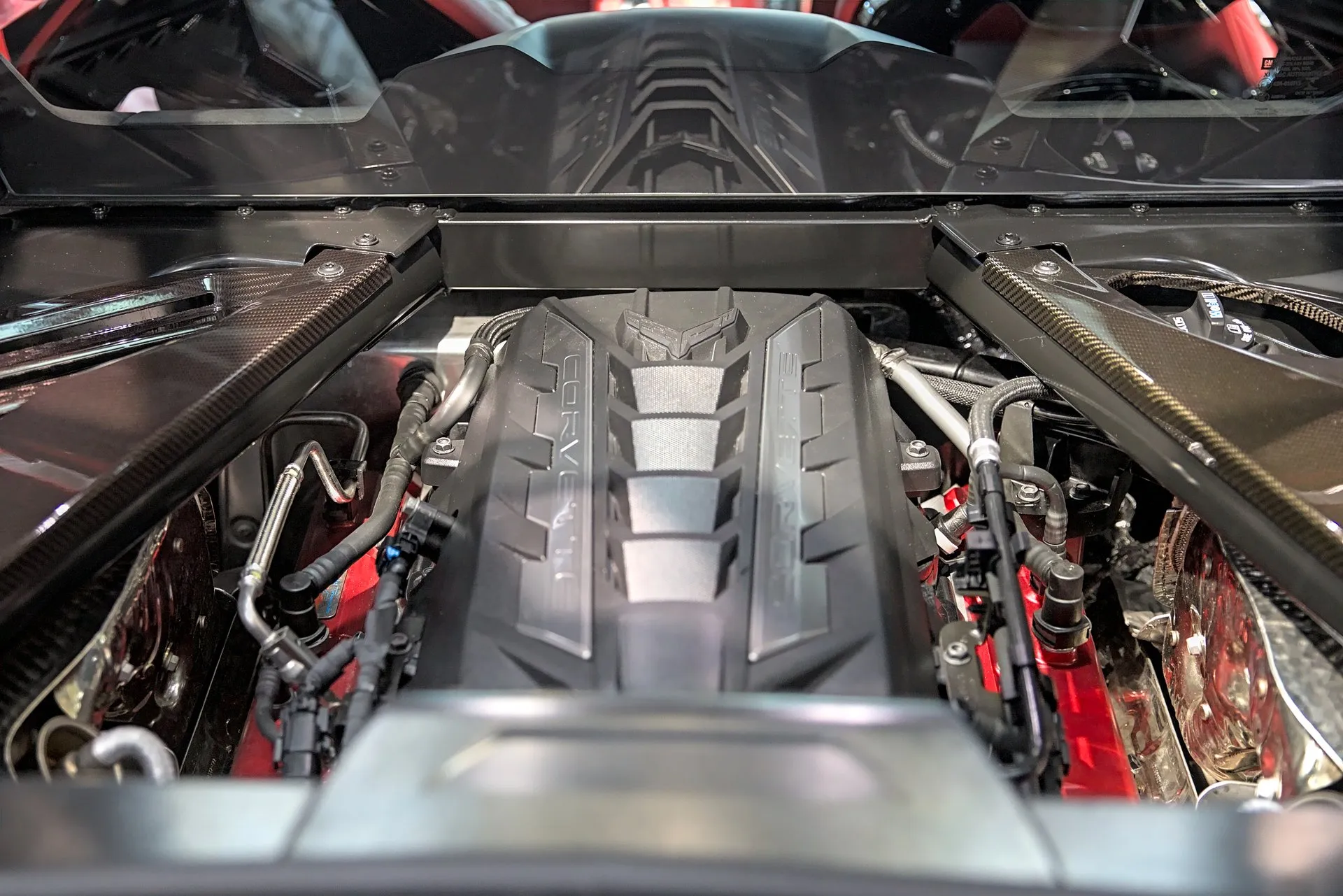 The top of a C8 Corvette's V8 under a black engine cover lettered Corvette, seen through the open rear hatch, with carbon-fiber braces on either side and the cabin's rear window beyond.
