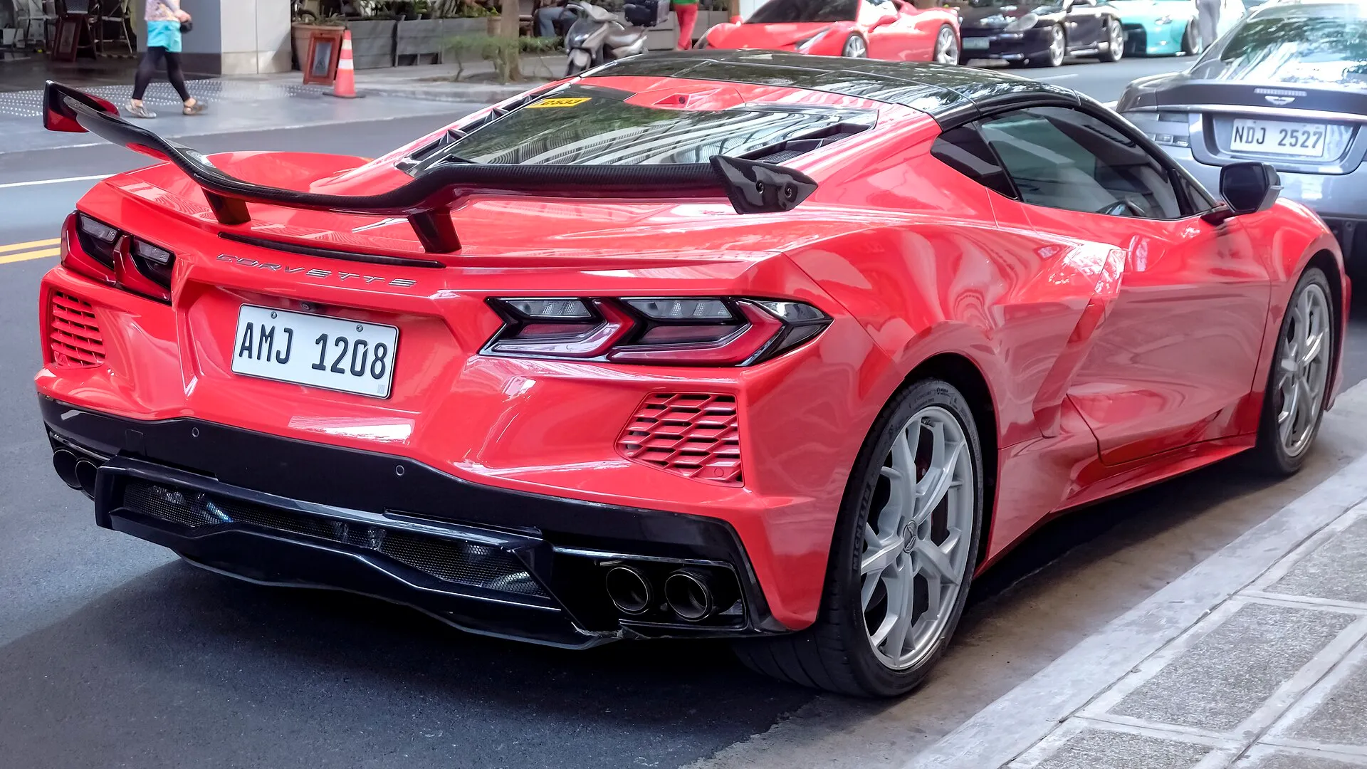 Red C8 Corvette Stingray coupe seen from the rear right corner, parked at a curb, with a tall black rear wing standing on two uprights above the engine cover.