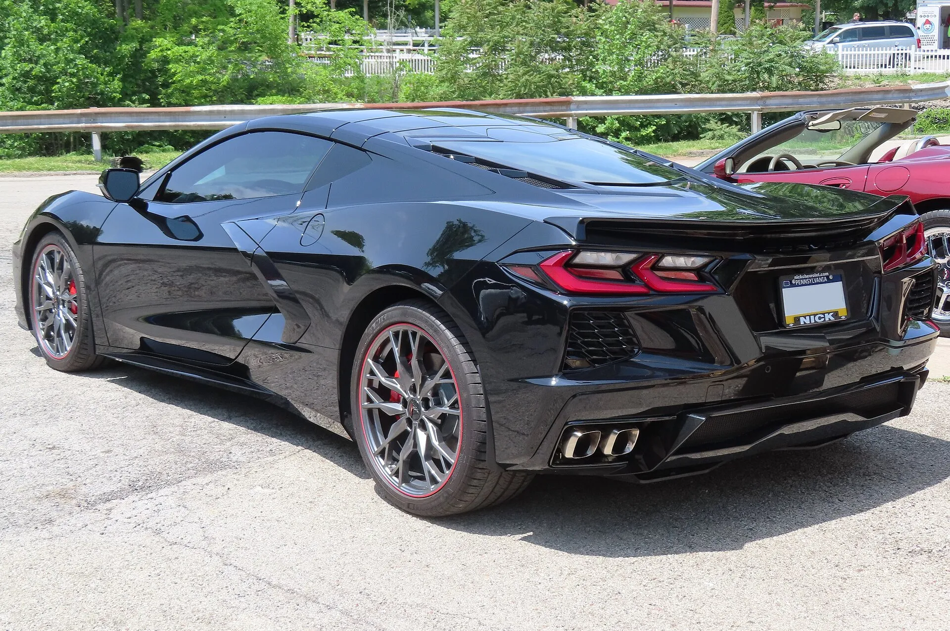 Black C8 Corvette Stingray coupe seen from the rear left corner in a parking lot, with a low lip spoiler across the tail and quad exhaust tips.