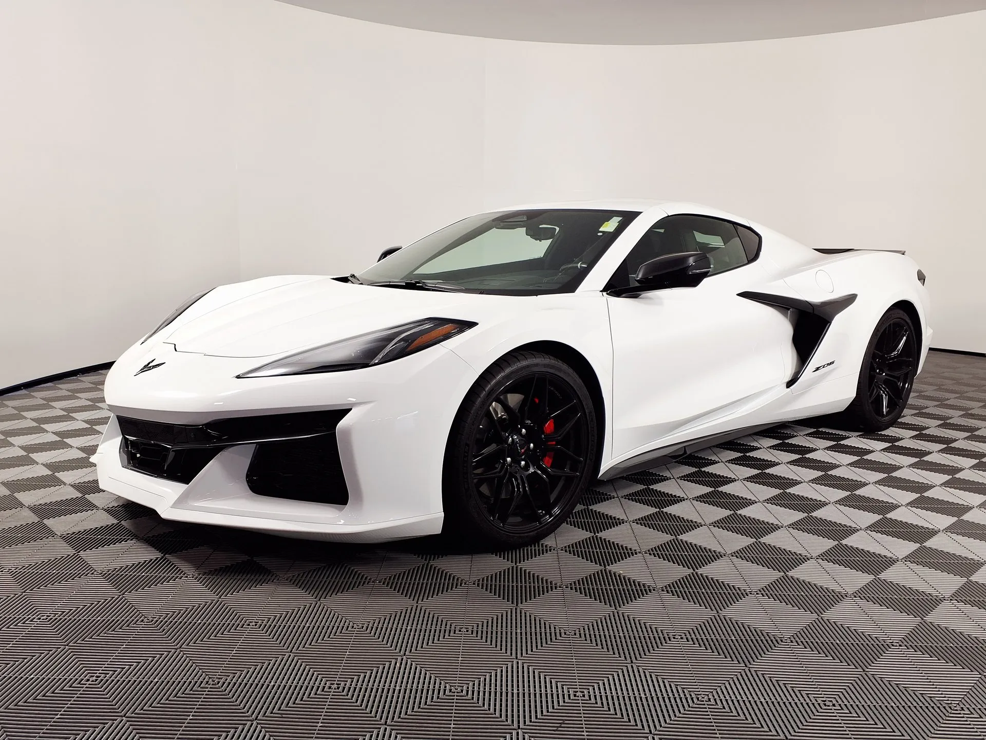 A white C8 Corvette Z06 coupe with black wheels, red brake calipers and black side air intakes, in a white photo studio with a checkered floor, seen from the front left.