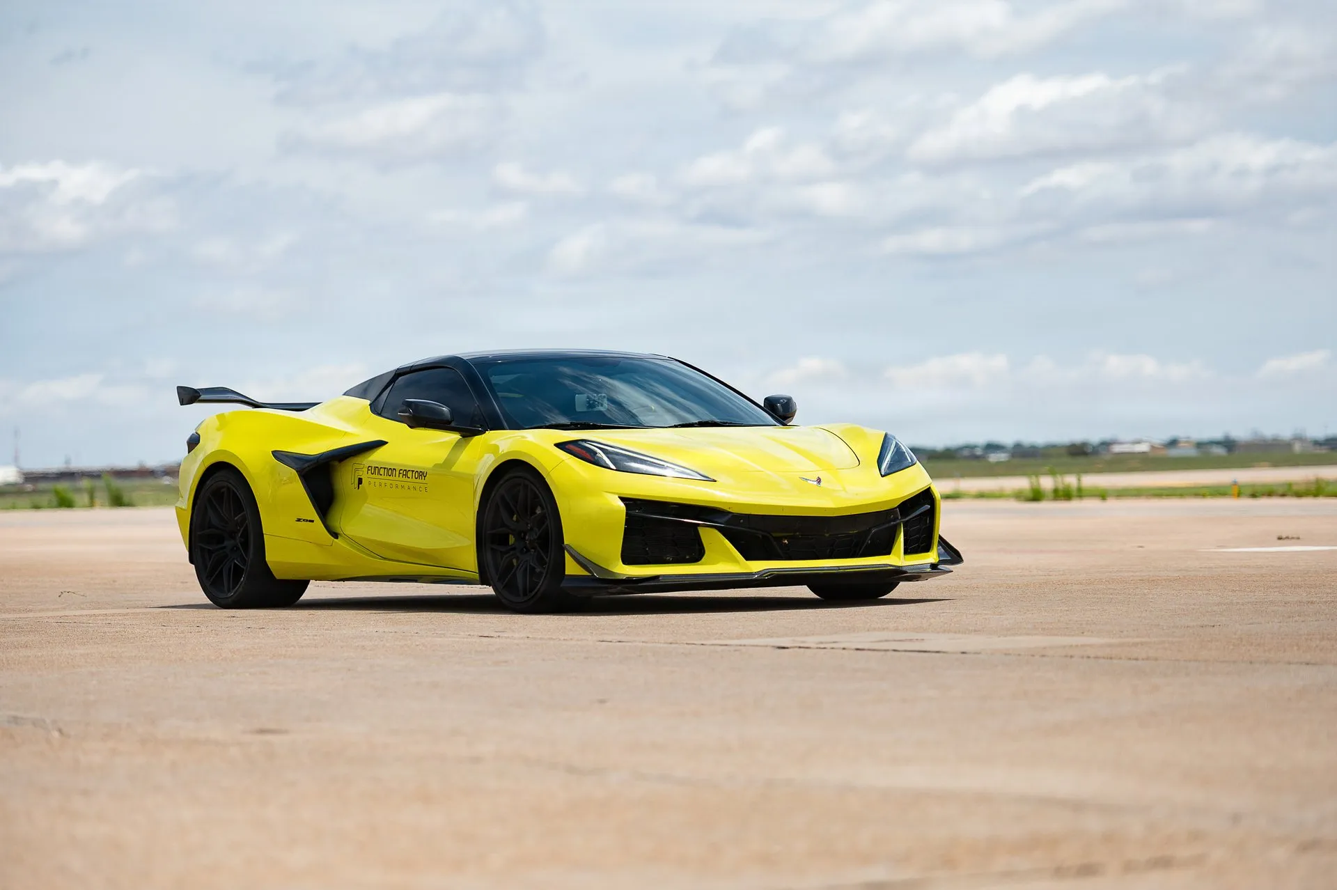 A yellow C8 Corvette Z06 coupe with a black roof, a tall black rear wing, black wheels, a carbon front splitter and a shop's decal on the door, on an airfield's concrete under a cloudy sky, seen from the front right.