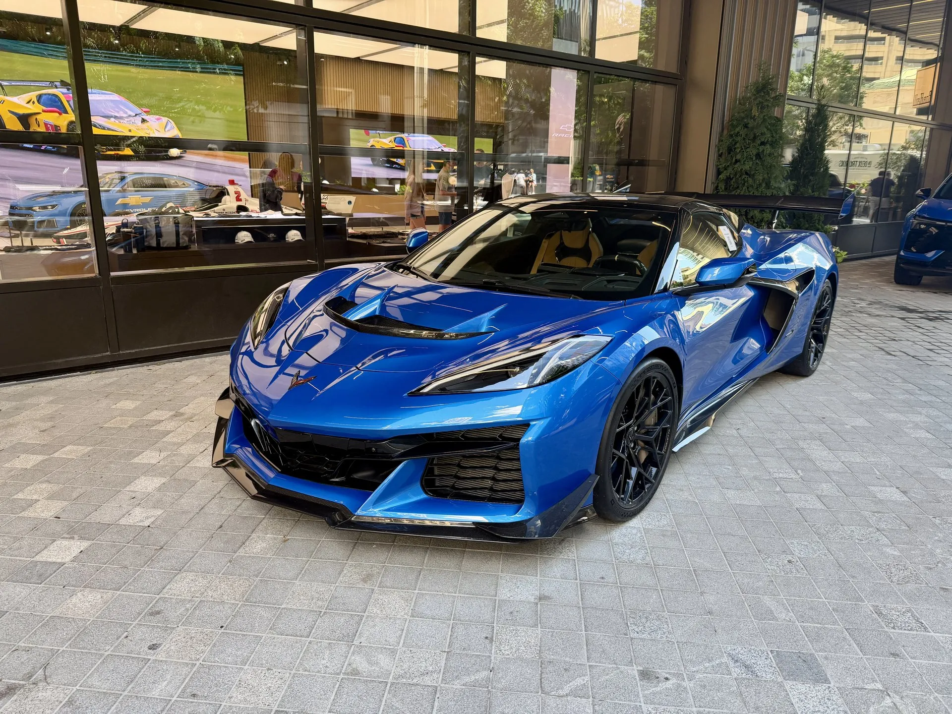 A blue C8 Corvette ZR1 coupe with a black roof, a vented hood, carbon-fiber trim and a tall rear wing, parked on stone pavers outside a glass-fronted building, seen from the front left.