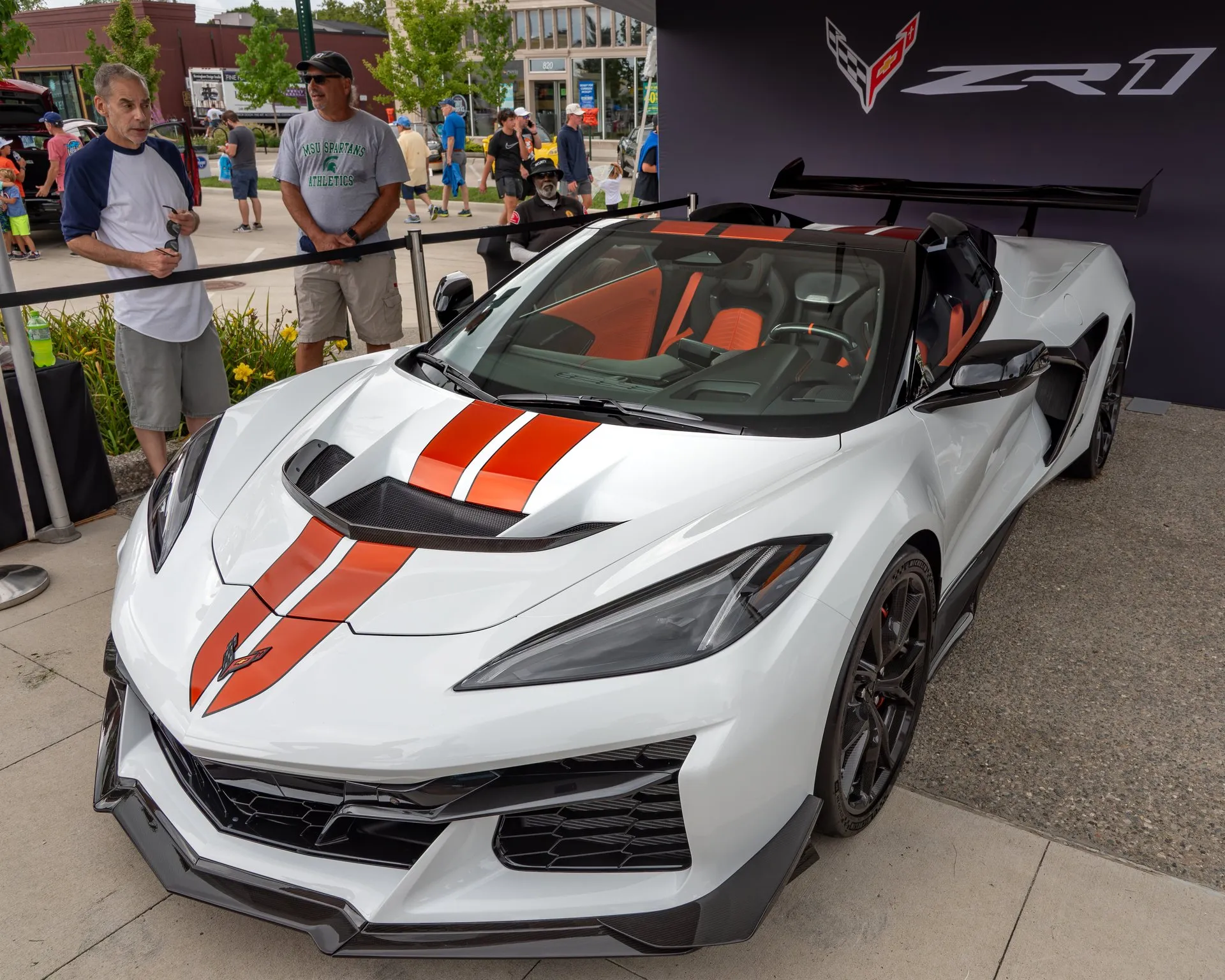A white C8 Corvette ZR1 convertible with its top stowed, twin orange stripes over the nose, a black vent through the hood, an orange and black interior, black wheels and a tall black rear wing, on display under a ZR1 banner at an outdoor car show, with visitors beside it.