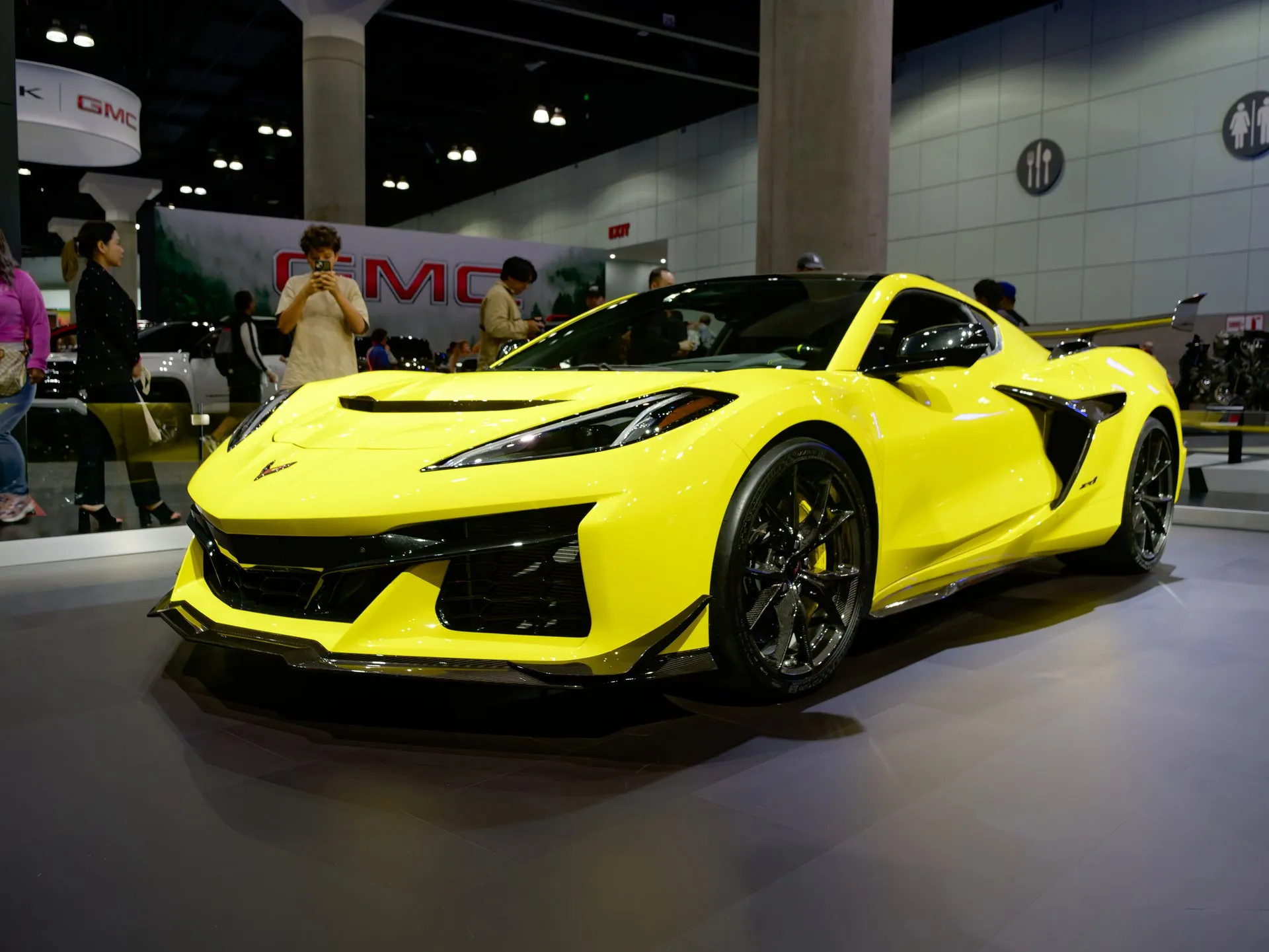 A bright yellow C8 Corvette ZR1 coupe on an auto show stand, seen from the front left: a black vent across the hood, a carbon-fiber front splitter, dark carbon-fiber wheels, yellow brake calipers and a tall rear wing, with show visitors behind it.