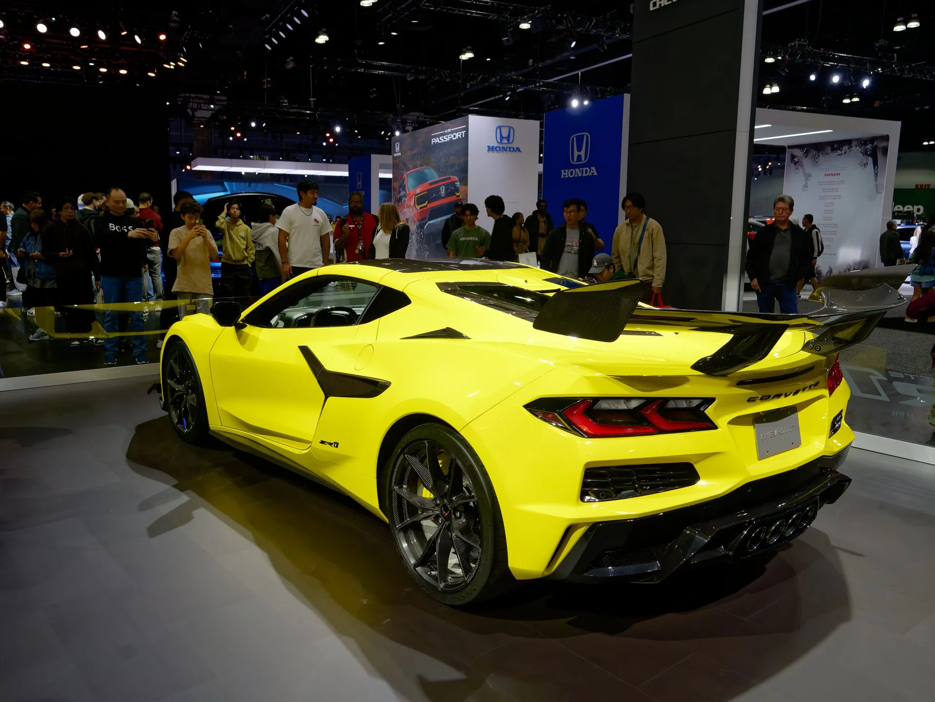 A yellow C8 Corvette ZR1 coupe seen from the left rear at an auto show: a tall carbon-fiber wing, four center exhaust tips, a ZR1 badge on the rear fender and carbon-fiber wheels, with a crowd of show visitors behind it.