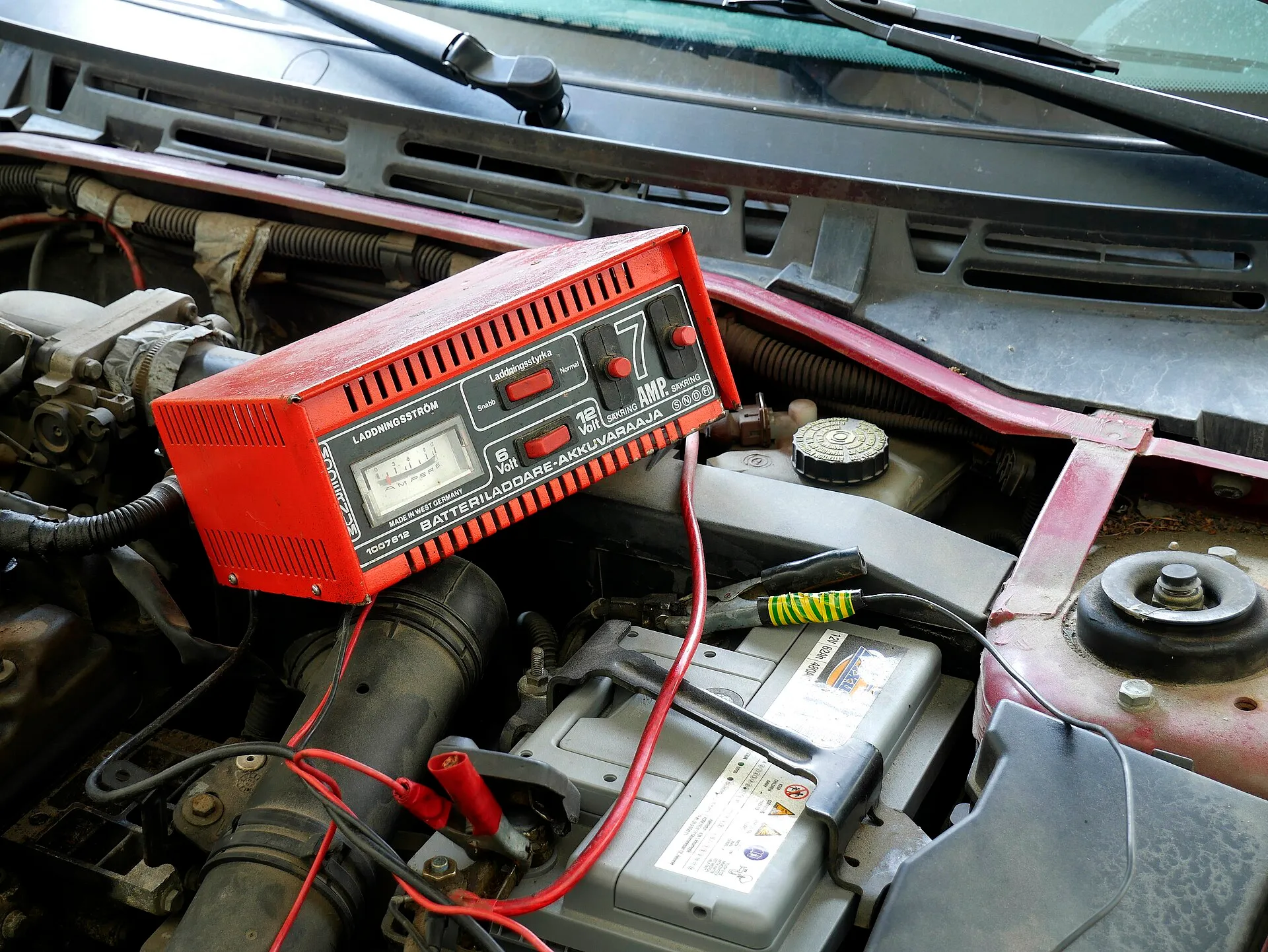 An old red manual battery charger with an ammeter and a charge-rate switch, sitting in an engine bay with its clamps on the car's battery.
