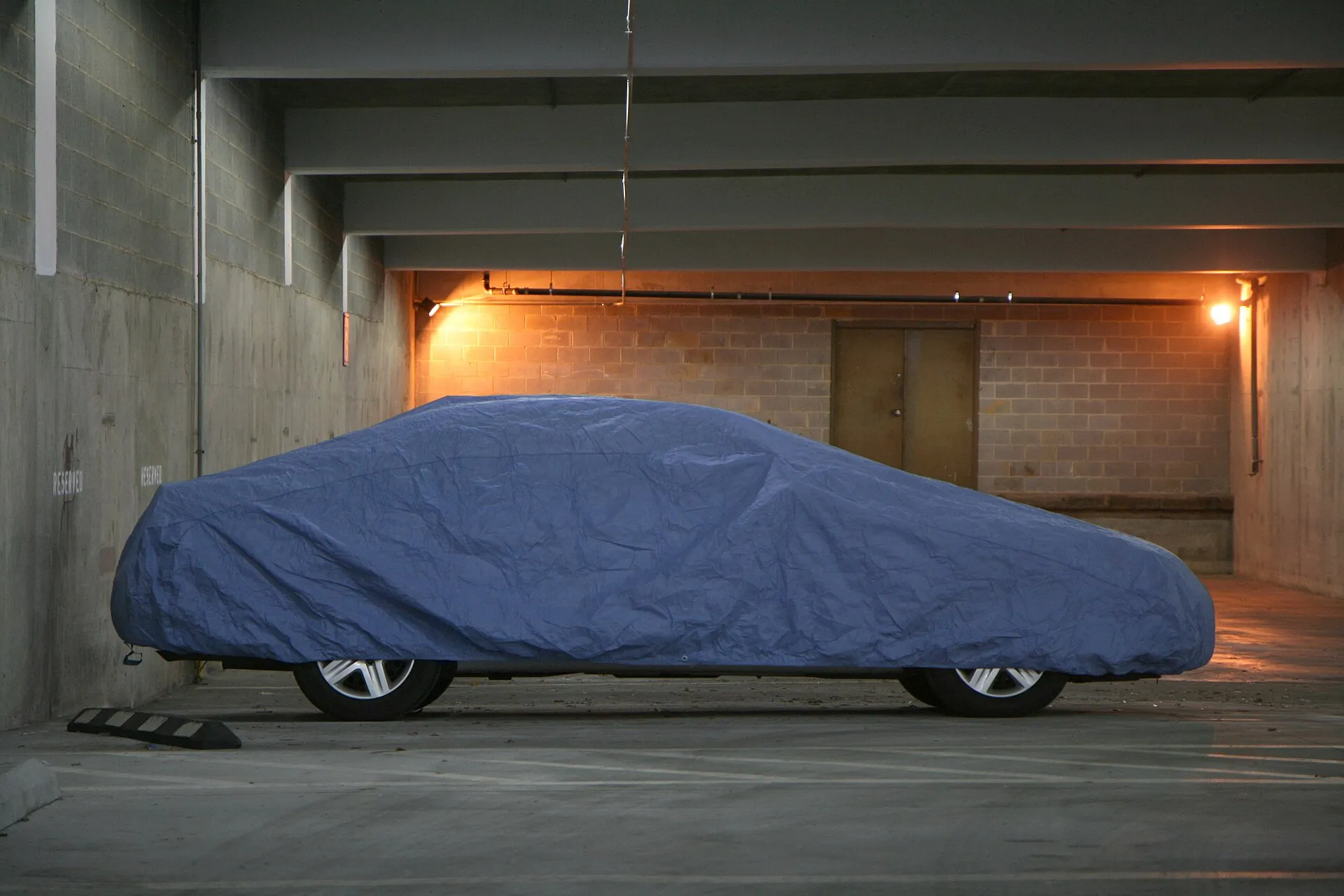 A car under a loose blue car cover in a concrete parking garage, the fabric wrinkled and slack along the sides.